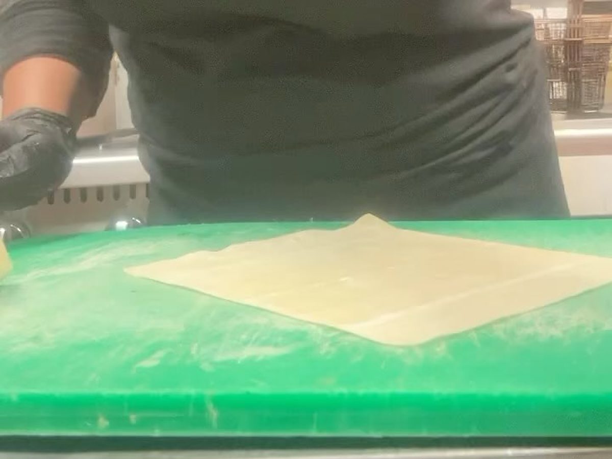 Daija at a stainless steel prep table in black gloves, rolling out an egg roll wrapper on a green cutting board. Fryer baskets sit on the shelf behind her.