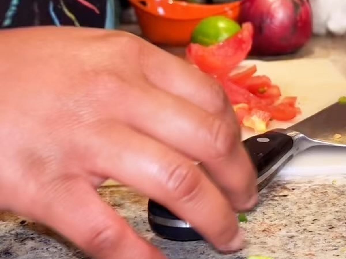 A hand steadying a chef knife through sliced tomato on a board, with a lime, a red onion and a small orange bowl beside it.